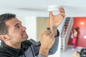 Ein Mann hält mit einer Hand einen Feuermelder an der Decke eines Raumes fest, in der anderen Hand hält er einen Schraubenzieher.